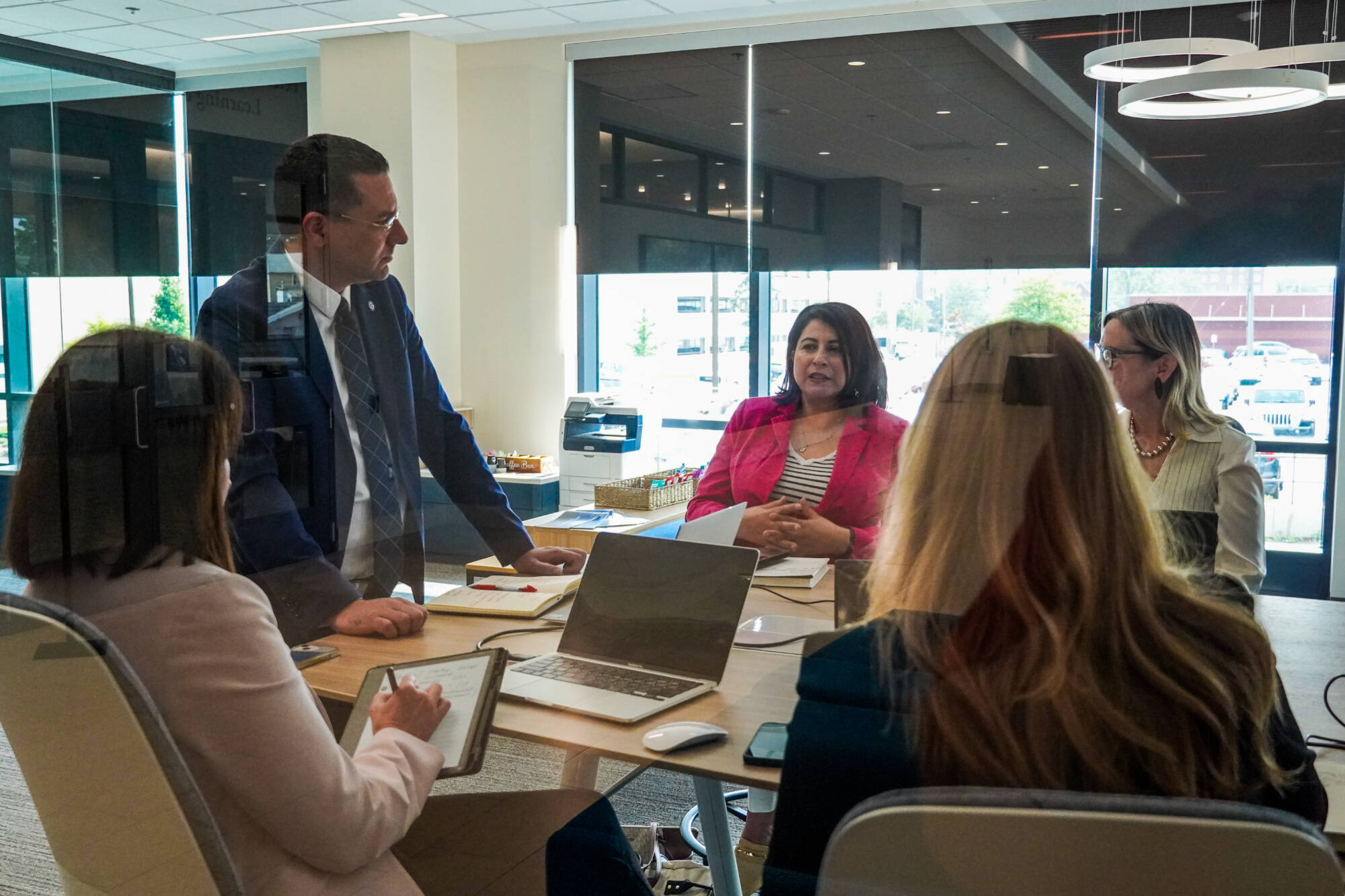 Five professionals gathered around a conference table in a modern glass-walled office, engaged in discussion. One man in a suit stands while four women, some with laptops and notebooks, are seated. Bright natural light fills the space from large windows.
